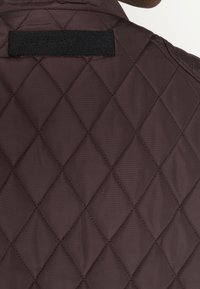 Giacca marrone trapuntata con etichetta logo nera ricamata al colletto. La texture superficiale presenta un motivo a diamante con una leggera lucentezza.