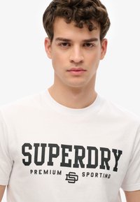 Hvid bomulds-T-shirt med en fed sort grafisk tryk, der står "SUPERDRY PREMIUM SPORTING." Klassisk rund hals design. Glat tekstur.