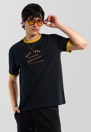 Młody mężczyzna w czarno-żółtej koszulce z napisem "GAS 1984 Authentic Denim Division", poprawiający bursztynowe okulary przeciwsłoneczne na jasnoszarym tle.
