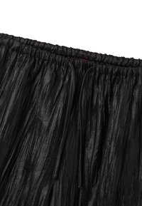 Falda negra plisada con cinturilla elástica. Está hecha de un material brillante y texturizado, con plisados largos y fluidos que permiten el movimiento.