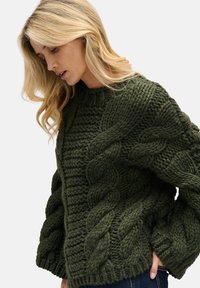 Ciemnozielony, grubo dziergany sweter z wzorem warkocza, teksturowanym ciałem i ściągaczem u dołu. Długie rękawy z wyraźnymi przeszyciami.