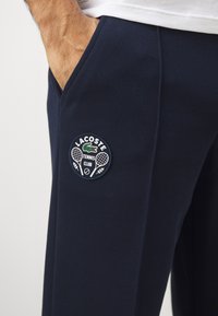 Sötétkék tenisznadrág, texturált anyagból, bal oldali zsebén a kerek Lacoste Teniszklub logóval.