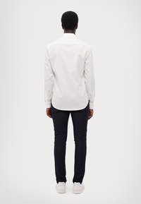 Λευκό πουκάμισο με μακριά μανίκια και απαλή υφή, συνδυασμένο με σκούρες στενές τζιν παντελόνες και λευκά αθλητικά παπούτσια, απεικονιζόμενο από πίσω σε φωτεινό φόντο.