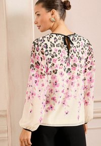 Blusa floreale con sfondo crema, caratterizzata da accenti maculati in rosa e nero. Maniche lunghe a sbuffo con dettaglio di scollo legato sulla schiena.