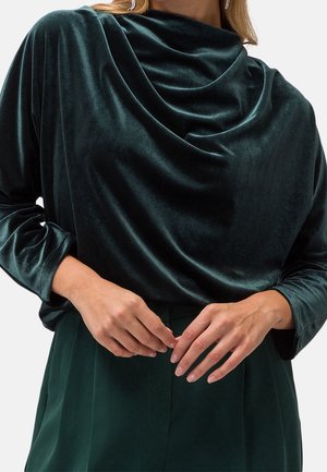 Femme portant un haut drapé en velours vert foncé à manches longues et un pantalon assorti vert foncé, les mains jointes près de la taille.