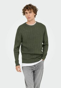 Pull en maille torsadée vert foncé avec un col rond. Tissu texturé avec des poignets et un ourlet côtelés. Superposé à une chemise blanche, porté avec un jean gris.
