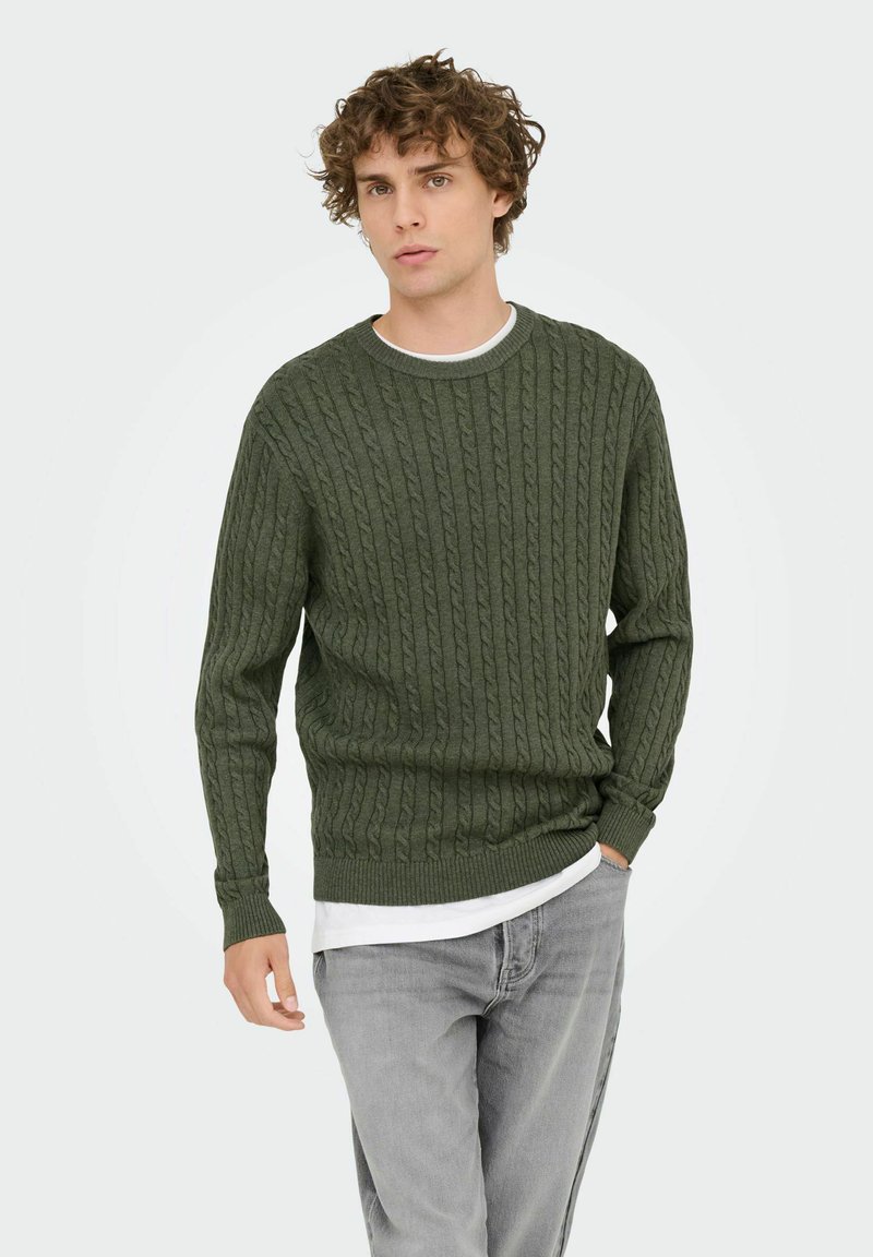 Pull en maille torsadée vert foncé avec un col rond. Tissu texturé avec des poignets et un ourlet côtelés. Superposé à une chemise blanche, porté avec un jean gris.