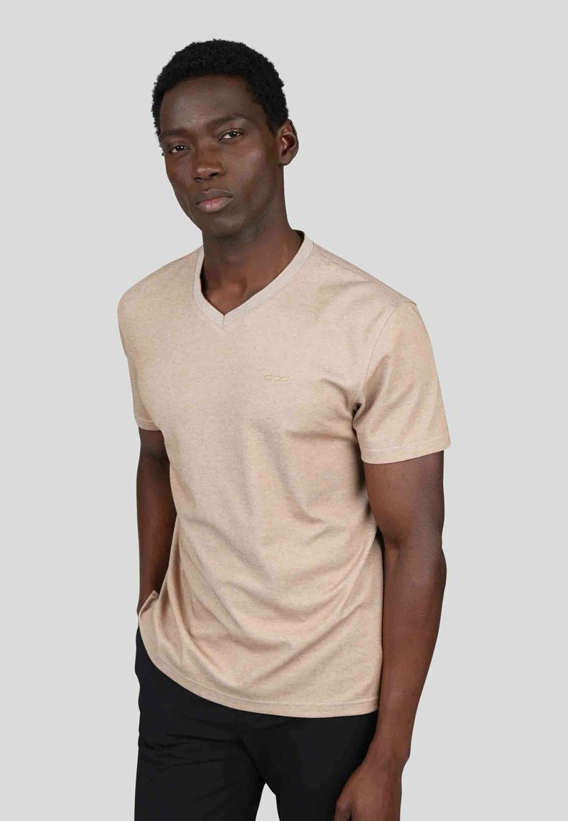 T-shirt en coton beige à col en V, à manches courtes, avec un logo subtil en or sur la poitrine et un tissu lisse légèrement texturé.