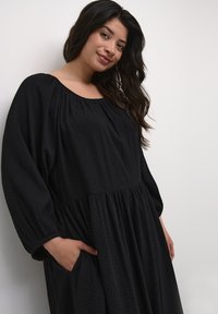 Black dress with a textured diamond pattern, loose fit, round neckline, and long sleeves. Features cinched waist and side pockets.