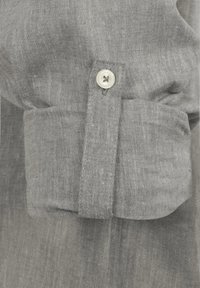 Puño de camisa de lino gris que presenta un solo botón redondo y una pestaña rectangular. La textura es ligeramente arrugada y transpirable.