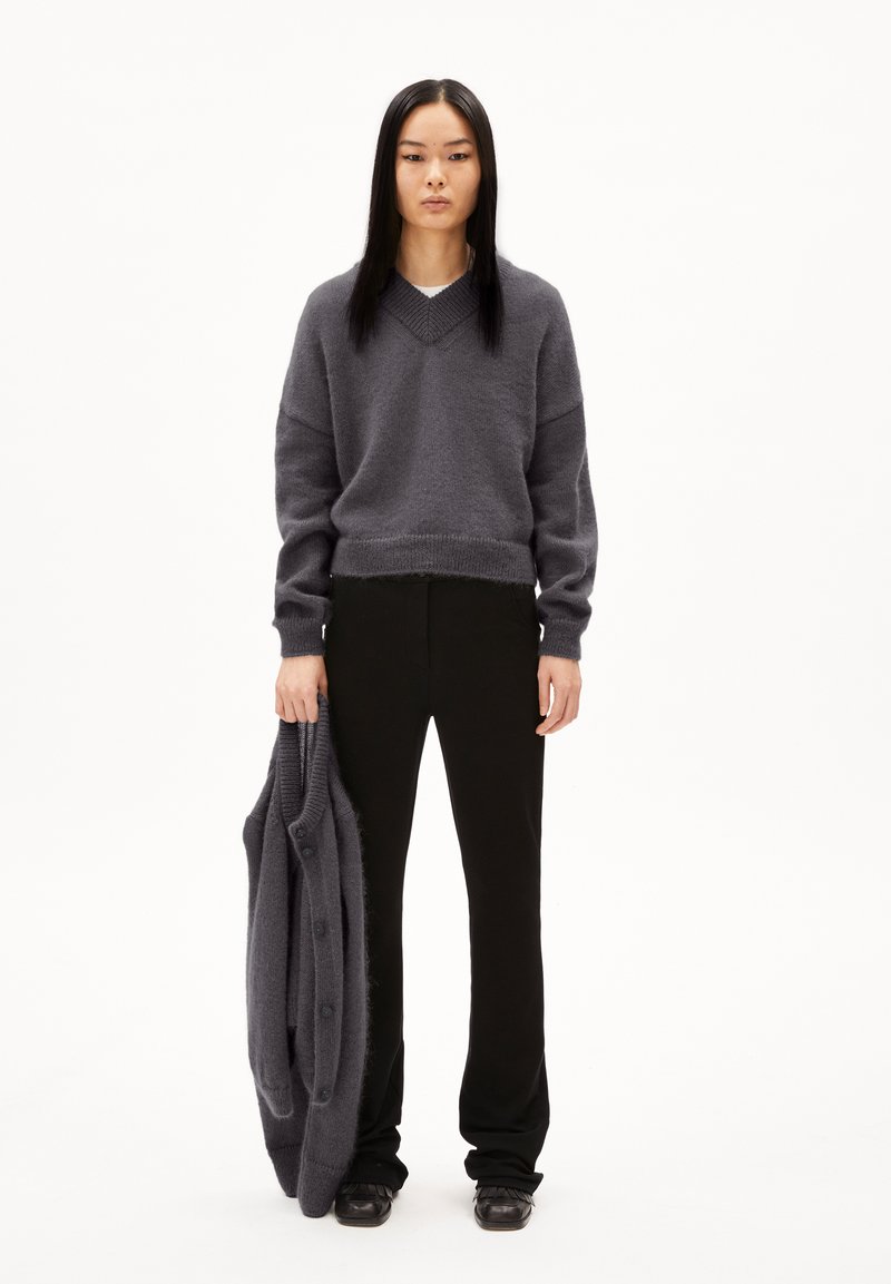 Grijze gebreide trui met een V-hals, gecombineerd met zwarte rechte broek. Model houdt een bijpassende grijze cardigan met knopen vast.