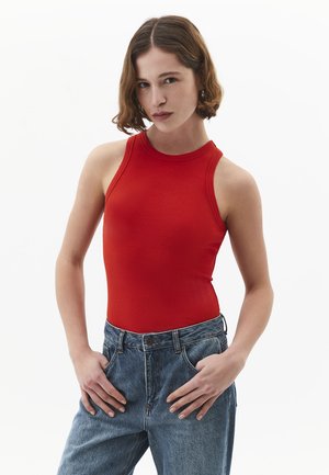 Giovane donna con capelli corti castani che indossa un top rosso senza maniche e jeans blu a vita alta, in piedi con le mani nelle tasche su uno sfondo bianco.