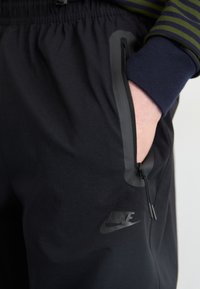 Černé sportovní kalhoty s bočními zipovými kapsami, elastickým pasem a logem Nike. Hladká látka s matným povrchem a elegantním designem.