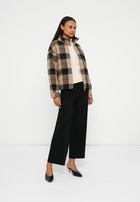 Beige, schwarze und braune karierte Jacke mit Frontreißverschluss und Stehkragen. Kombiniert mit weit geschnittenen schwarzen Hosen und schwarzen High Heels.
