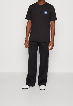 Czarna koszulka z kolorową okrągłą grafiką na lewej piersi, w połączeniu z czarnymi spodniami z białymi paskami po bokach i białymi sneakersami.