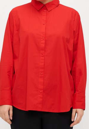 Persona con una camicia a maniche lunghe di un vivace rosso, con colletto e bottoni, abbinata a pantaloni neri, su uno sfondo chiaro e neutro.