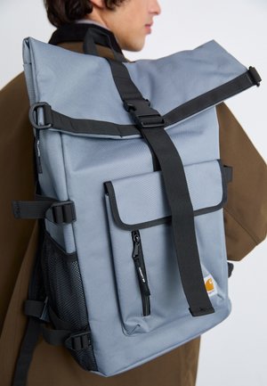 Person wearing a brown coat carrying a large light grey backpack with black straps and a front zippered pocket.