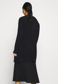 Cárdigan largo de marina confeccionado en una textura suave y esponjosa, con un corte relajado y aberturas laterales, combinado con una falda negra con pliegues.