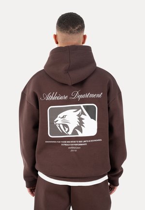 Personne portant un sweat à capuche marron avec un logo de panthère rugissante blanche et le texte "Athleisure Department" ainsi qu'un slogan motivant au dos.