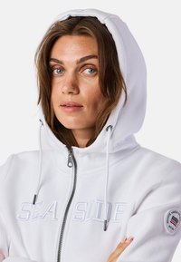 Weißer Reißverschluss-Hoodie mit Kapuze, ausgestattet mit aufgesticktem Schriftzug "SEASIDE" und einem runden Logo-Patch auf dem linken Ärmel. Softer Stofftextur.