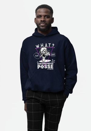 Mężczyzna noszący granatową bluzę z kapturem z grafiką kreskówkowej szczura w kasku oraz tekstem "W.H.A.T? Subterranean Posse Since 1967," w połączeniu z czarnymi, kraciastymi spodniami.