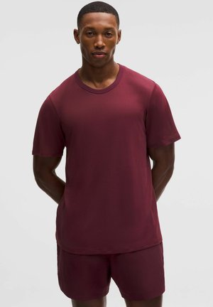 Mann steht mit den Händen hinter dem Rücken, trägt ein passendes dunkelrotes kurzärmeliges Hemd und Shorts vor einem schlichten, hellen Hintergrund.