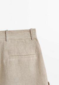 Pantalon en lin beige montrant la zone de la taille avec passants de ceinture et une seule poche arrière horizontale sur fond blanc.