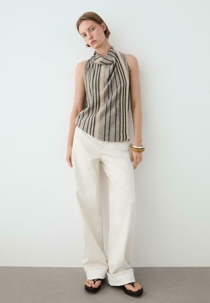 Frau trägt ärmelloses gestreiftes beiges und schwarzes Oberteil, weite weiße Hose, schwarze Sandalen und Holzarmbänder, steht an einer weißen Wand.