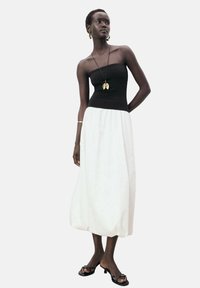 Schwarzes trägerloses Oberteil kombiniert mit einem fließenden weißen Maxirock. Die Accessoires umfassen eine goldene Halskette und schlichte schwarze Sandalen. Glatte Stofftextur.