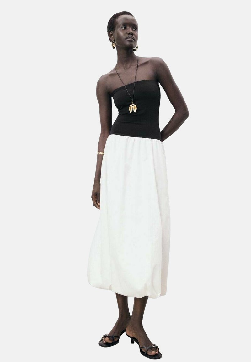 Schwarzes trägerloses Oberteil kombiniert mit einem fließenden weißen Maxirock. Die Accessoires umfassen eine goldene Halskette und schlichte schwarze Sandalen. Glatte Stofftextur.