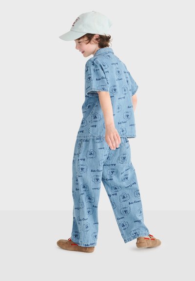 Enfant portant une chemise en jean bleu clair et un pantalon avec des dessins animés, une casquette beige et des chaussures marron, marchant de côté sur un fond uni.