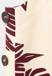 Tissu crème avec un motif abstrait de feuilles rouge foncé, avec deux grands boutons foncés cousus sur le bord.