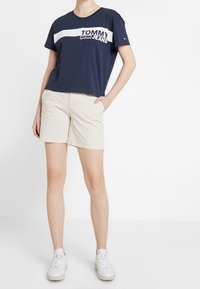 Marinblå bomullströja med vit "TOMMY JEANS"-logotyp, i kombination med beige shorts och vita sneakers. T-shirten har en avslappnad passform och rund halsringning.