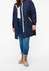 Marineblå quiltet lang frakke med cremet lynlås, sidelommer og ribbede manchetter. Bæres over en lys top og blå jeans, parret med lysebrune snørebåndsstøvler.