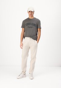 Camiseta gris de manga corta con estampado negro "TOMMY JEANS New York", combinada con pantalones color crema y zapatillas blancas. Gorra blanca en la cabeza del modelo.