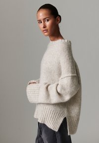 Camisola de lã cinzento claro com decote barco, corte oversized, punhos canelados e fendas laterais. Textura macia com um acabamento ligeiramente felpudo.