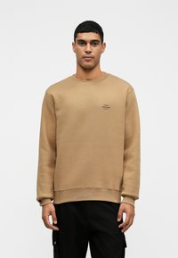 Férfi bézs színű kereknyakú pulóverben, amely bal mellrészen egy apró fekete logóval van ellátva, fekete nadrágban, sima fehér háttér előtt áll.