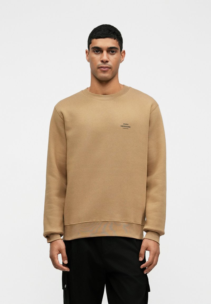 Férfi bézs színű kereknyakú pulóverben, amely bal mellrészen egy apró fekete logóval van ellátva, fekete nadrágban, sima fehér háttér előtt áll.