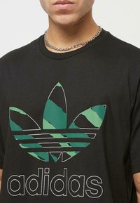 Zwarte katoenen T-shirt met een groot groen en zwart Adidas-logo, inclusief drie bladvormen en twee horizontale strepen eronder.