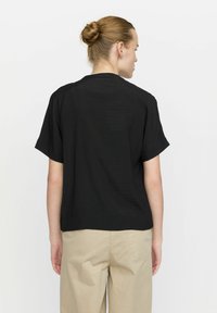 Camicetta nera a maniche corte con vestibilità rilassata, caratterizzata da una superficie testurizzata. Abbinata a pantaloni beige chiaro. La vista posteriore mostra un design minimalista.