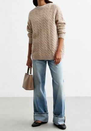 Personne portant un pull beige en maille torsadée, un jean bleu clair à jambes larges avec revers, des ballerines noires, et tenant un sac à main beige avec des poignées foncées.