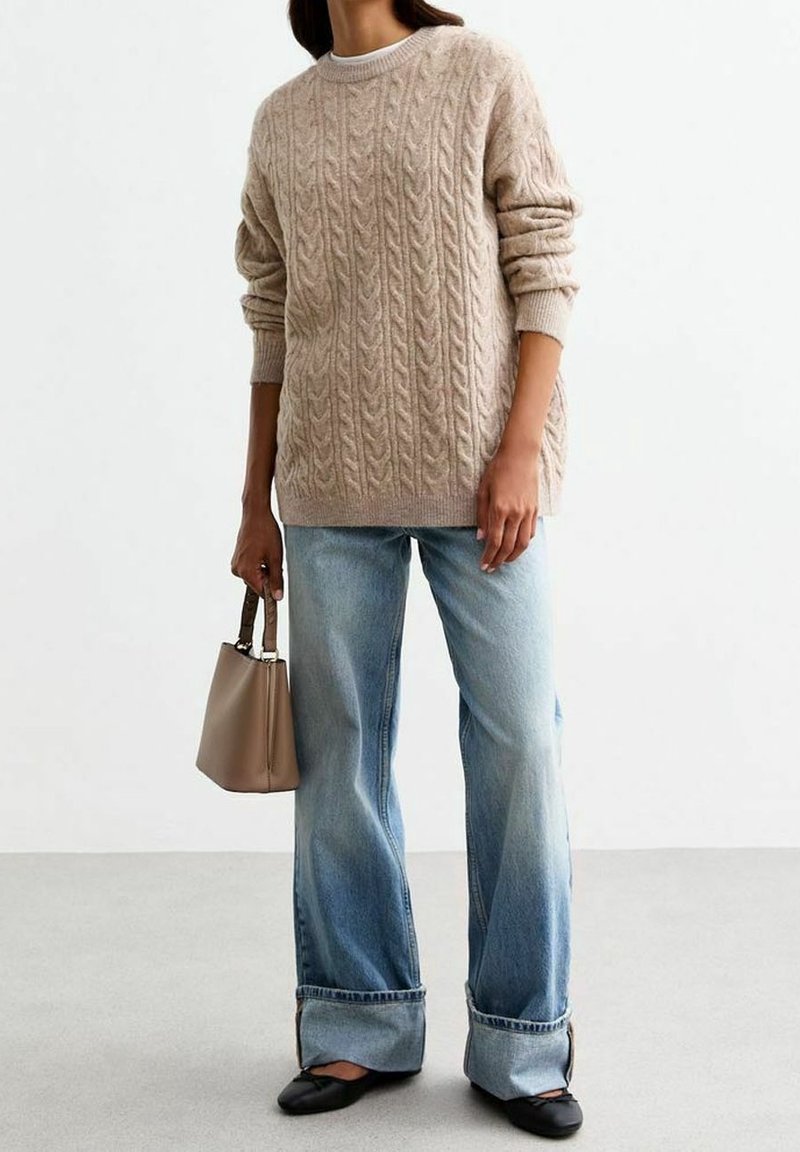 Personne portant un pull beige en maille torsadée, un jean bleu clair à jambes larges avec revers, des ballerines noires, et tenant un sac à main beige avec des poignées foncées.