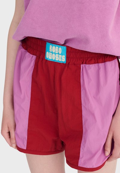 Personne portant un short rouge et rose avec une ceinture élastique et une étiquette bleue indiquant "Bobo Choses", associé à un haut violet.