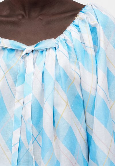 Άτομο με σκουρόχρωμο δέρμα που φοράει ρούχο ανοιχτού γαλάζιου και λευκού καρό με δεμένο φιόγκο στο ντεκολτέ, που δείχνει το πάνω μέρος του στήθους και τον ώμο.