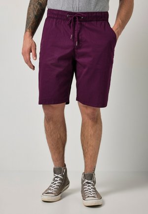 Mann trägt lila Kordelzug-Shorts mit Taschen und graue High-Top-Sneaker und steht vor einem einfachen Hintergrund.