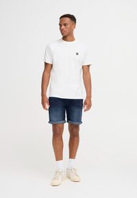 Fehér rövid ujjú póló fekete hatszög alakú folttal, sötétkék farmer rövidnadrággal, amelynek szegélye fel van hajtva, valamint világos bézs színű tornacipővel.