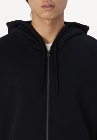 Sudadera negra con cremallera, textura suave, capucha con cordón y diseño minimalista con una sutil marca en el pecho.