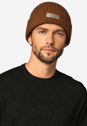 Uomo con la barba che indossa un berretto marrone a coste con l'etichetta "Bellemere New York" e un maglione girocollo nero, rivolto in avanti su uno sfondo bianco.