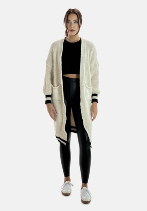 Joven mujer de pie, con un cárdigan largo beige de punto sobre un crop top negro y leggings negros con zapatillas blancas, fondo neutro.