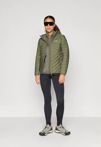 Kvinne står iført mørke solbriller, olivengrønn pufferjakke, mørke tights og grå turstøvler mot en ensfarget bakgrunn.